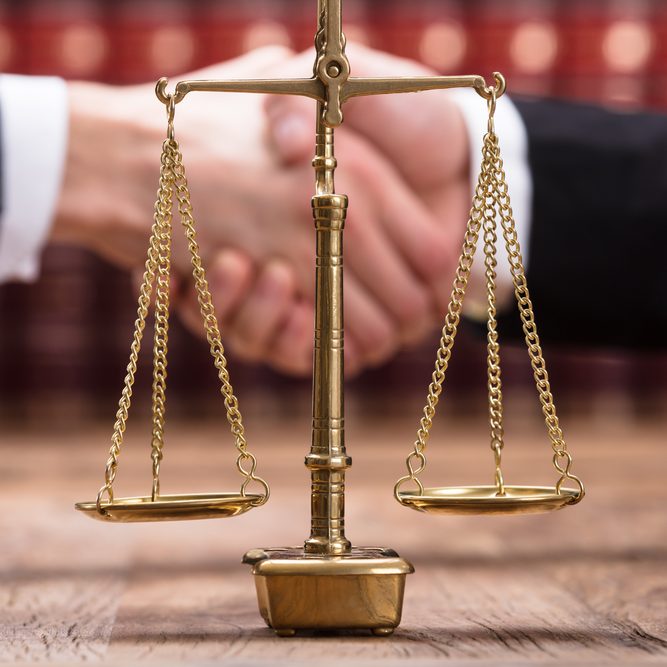 Close-up Of Justice Scale On Wooden Desk In Front Of Businesspeople Shaking Hands